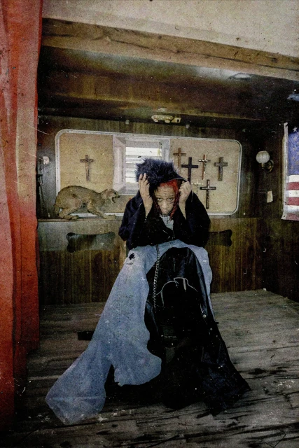 Woman with dark hair sits indoors in front of religious display, with an American flag visible, wearing black coat and blue pants.