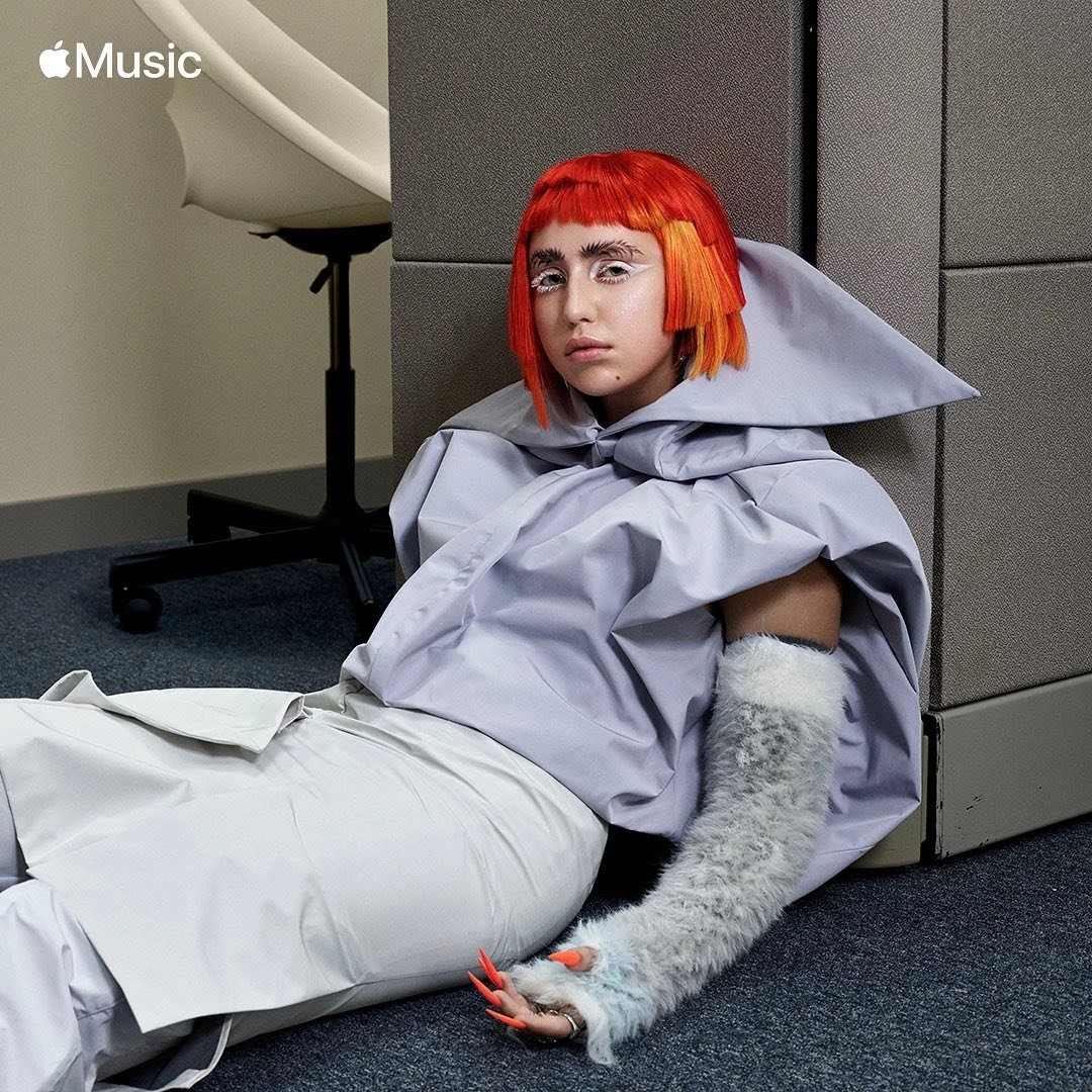 Portrait of a person with bright orange hair and white makeup sitting on the floor near a gray wall and an office chair.