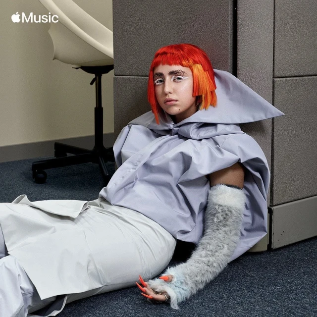 Portrait of a person with bright orange hair and white makeup sitting on the floor near a gray wall and an office chair.