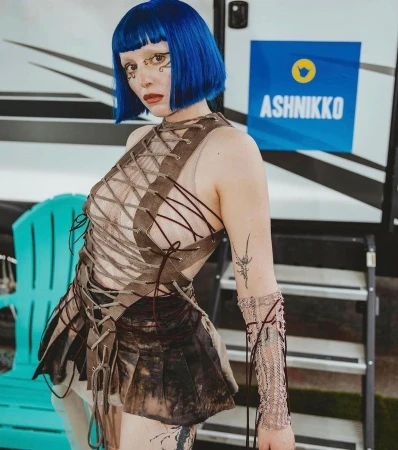 A woman with bright blue hair, face makeup, and a unique laced outfit stands in front of a travel trailer with an 'Ashnikko' logo.