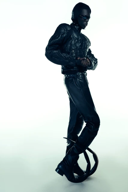 Fashionable adult in black leather jacket and pants poses indoors on avant-garde prop with a high-key, high-contrast studio lighting.