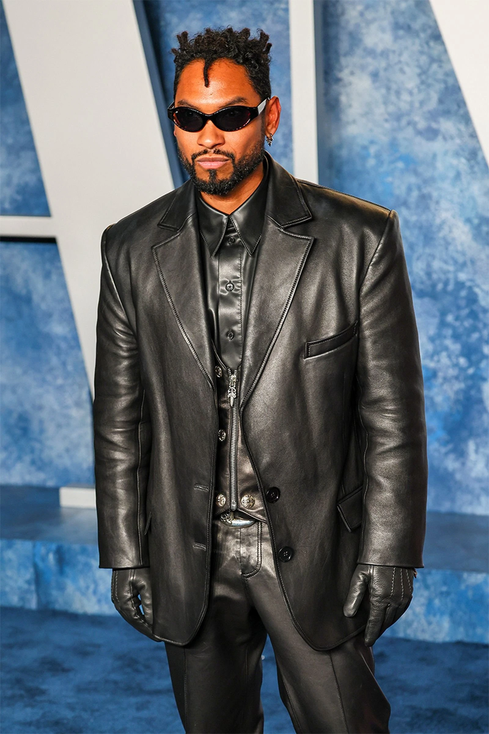 A well-dressed Black man in a black leather suit and tinted glasses poses against a faded blue backdrop for a celebrity event.