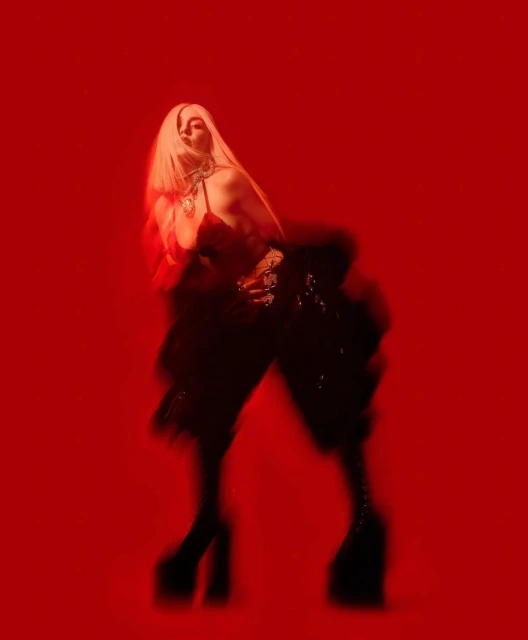 A woman with platinum blonde hair poses in a red-toned photo, wearing a dark outfit and platform boots against a solid red background.
