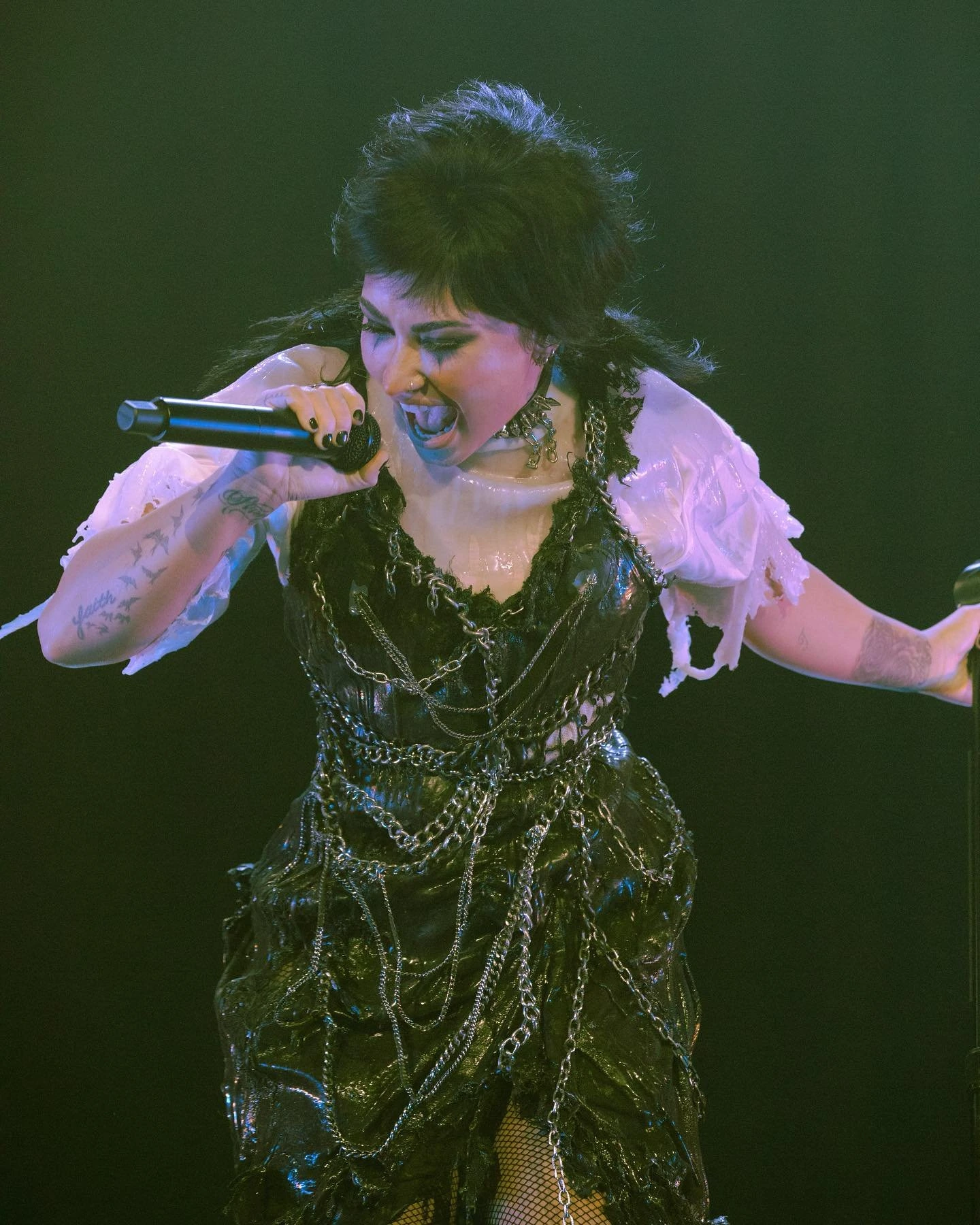 Adult female vocalist with dark hair and elaborate costume singing with a microphone on a dark stage with dramatic lighting during performance.