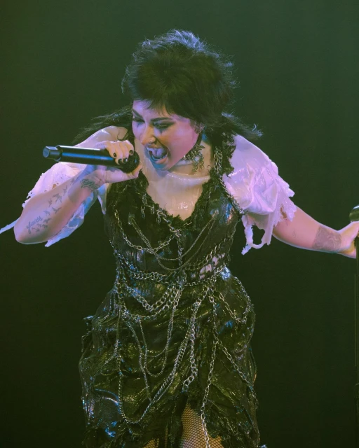 Adult female vocalist with dark hair and elaborate costume singing with a microphone on a dark stage with dramatic lighting during performance.