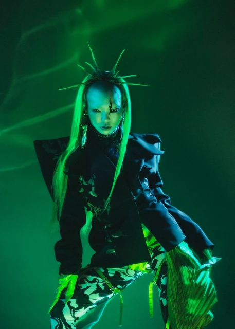 Woman with green hair poses in black patterned outfit against a green background with light effects, creating a striking fashion editorial image.