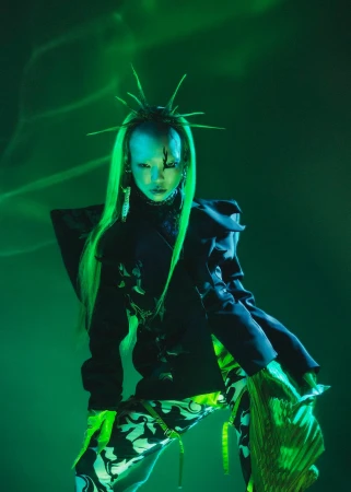 Woman with green hair poses in black patterned outfit against a green background with light effects, creating a striking fashion editorial image.