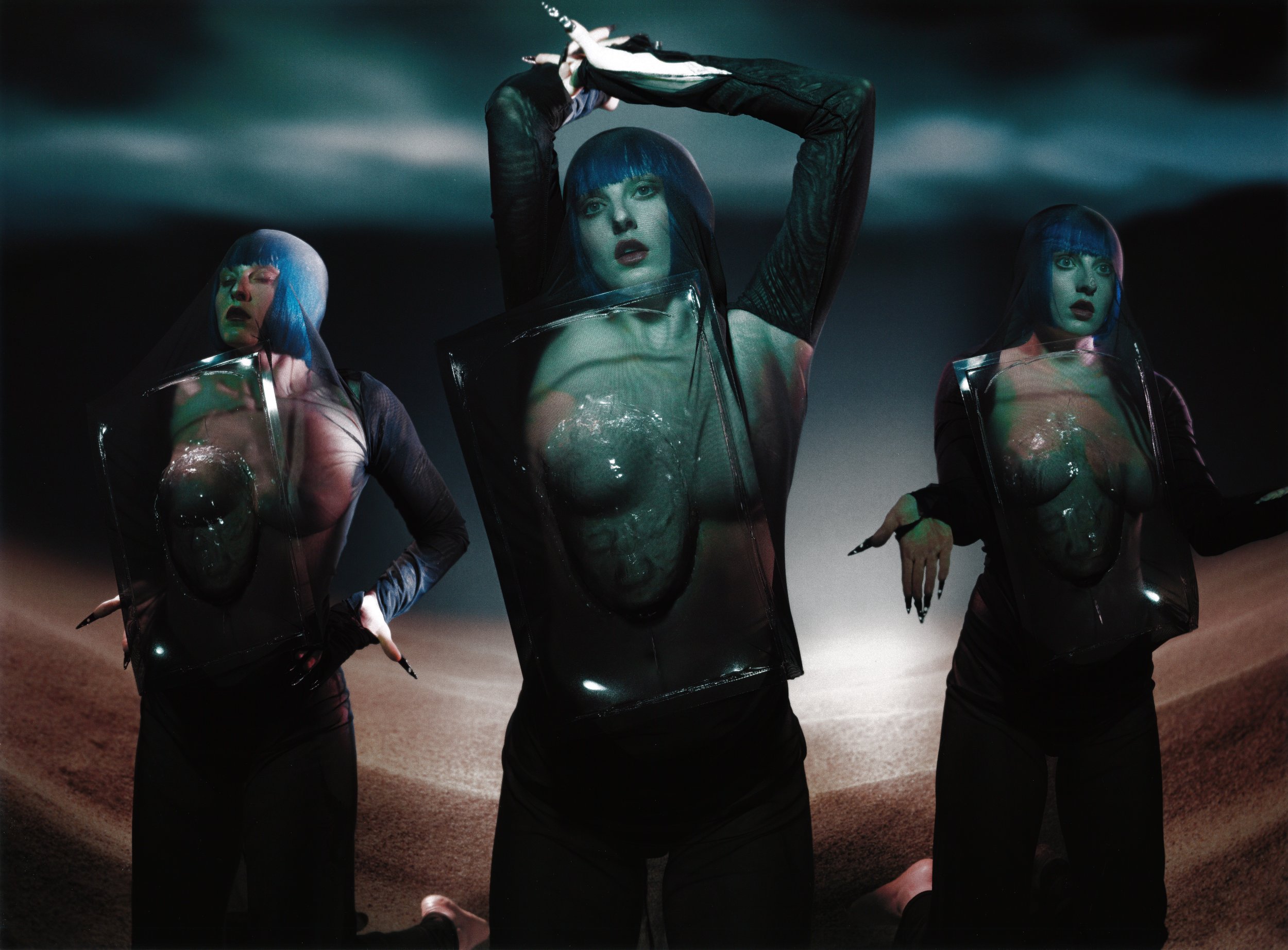 Three women with blue wigs and unique clothing pose in an artistic photo. The scene features a surreal, staged setting with dramatic lighting.