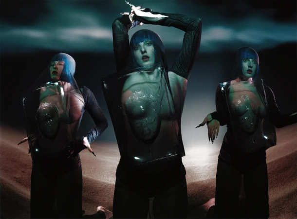 Three women with blue wigs and unique clothing pose in an artistic photo. The scene features a surreal, staged setting with dramatic lighting.
