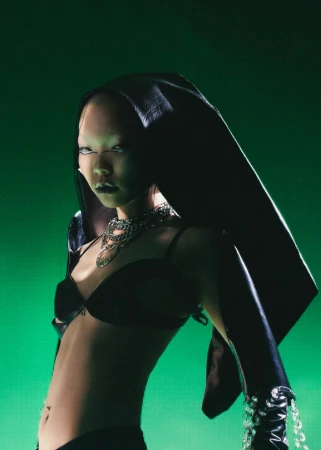 Model wearing a black bra, head covering, and layered silver chain necklaces against a moody green background.