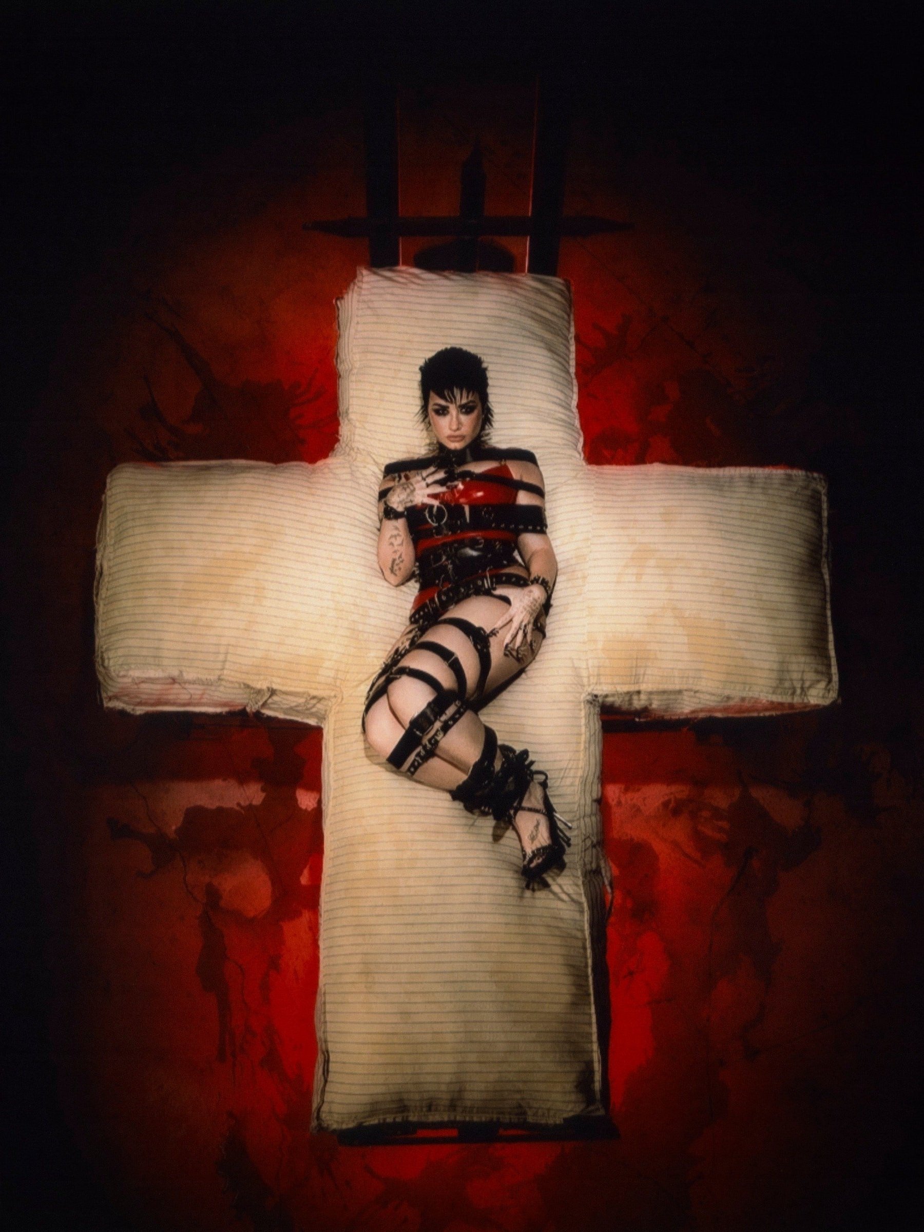 Woman in platform heels and a black and red corset poses dramatically on a cross-shaped bed in a dark, smoky music studio setting.