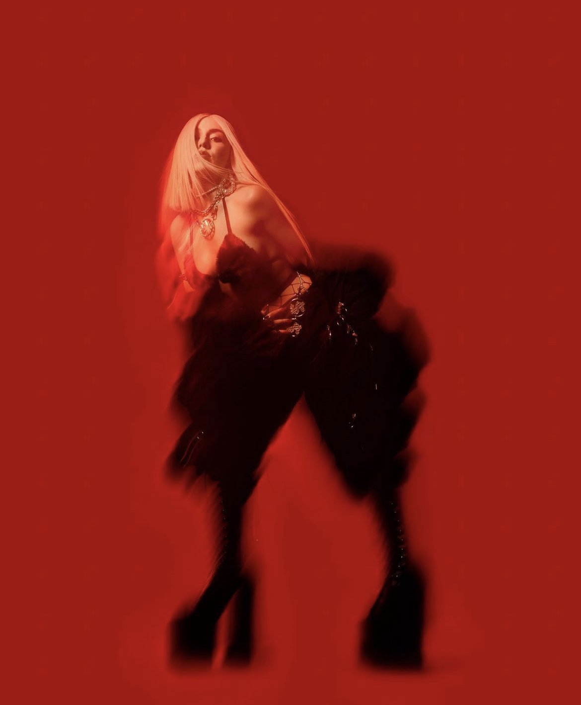 Fashionable adult female posing in a red studio wearing black platform boots, oversized pants, a black corset top and silver jewelry.