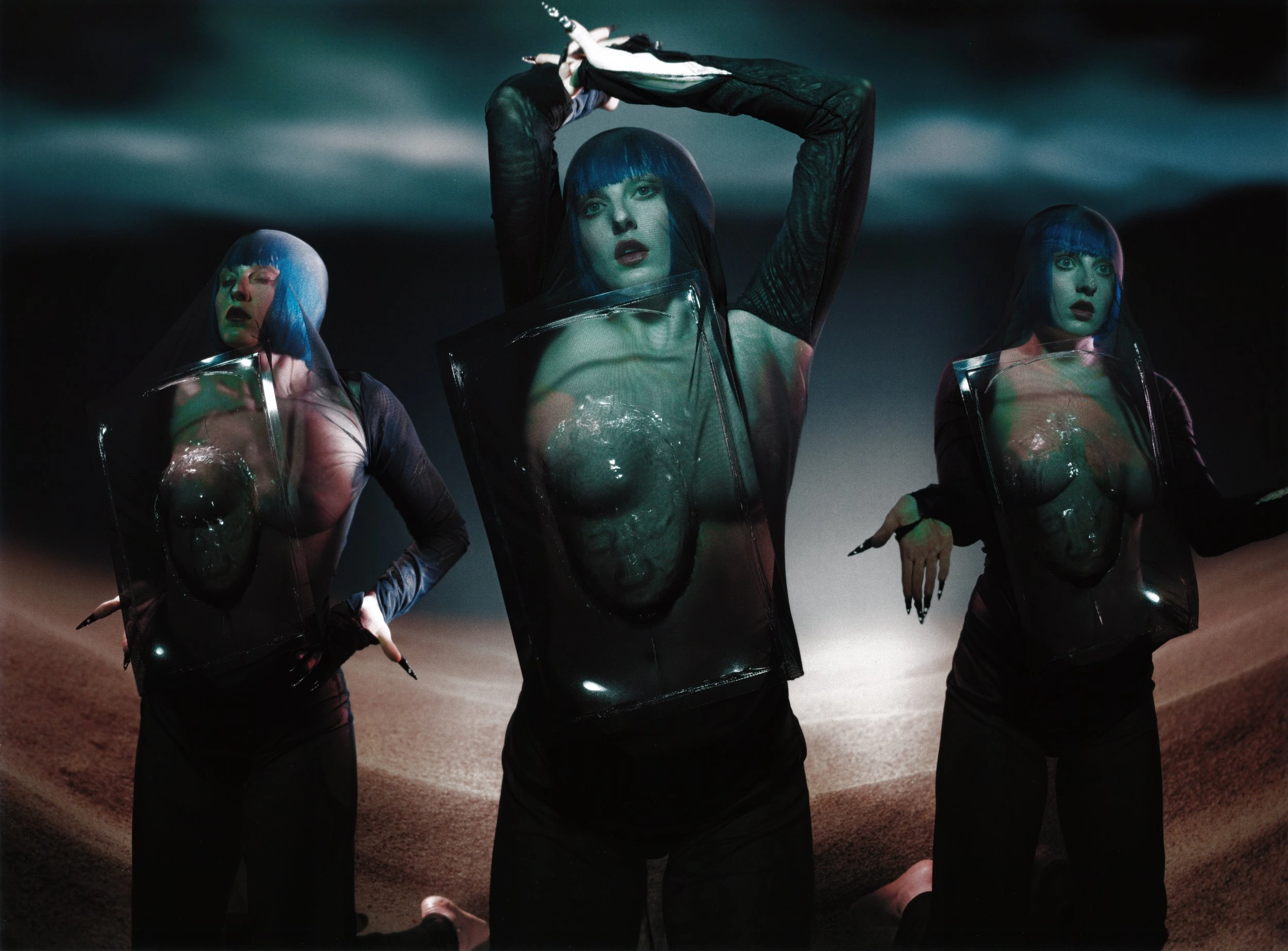 Three figures with blue hair and dark clothing pose against a dark background with ground, creating a surreal and futuristic aesthetic.