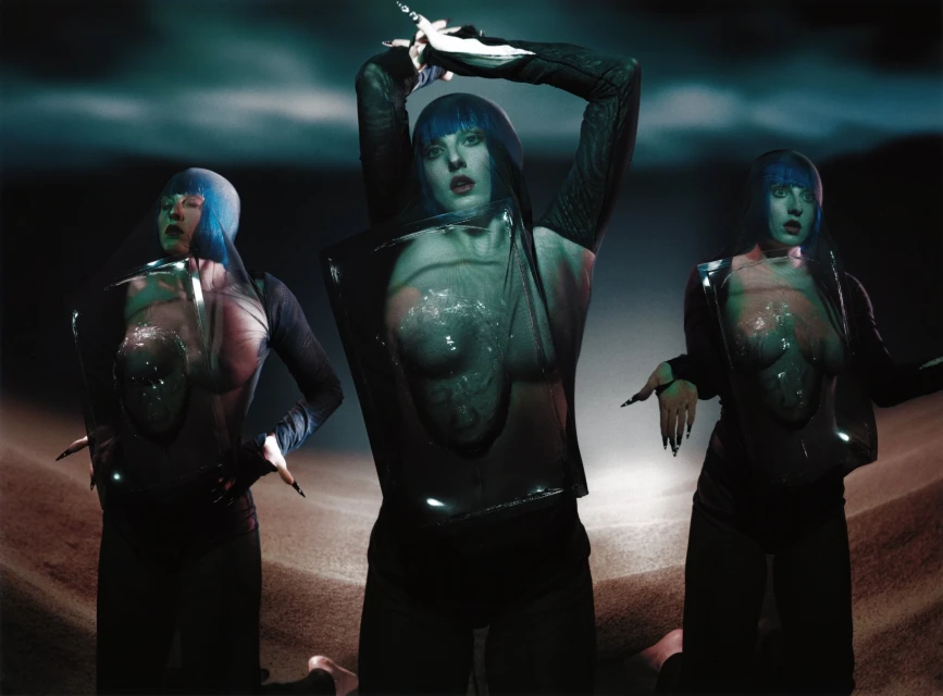 Three figures with blue hair and dark clothing pose against a dark background with ground, creating a surreal and futuristic aesthetic.