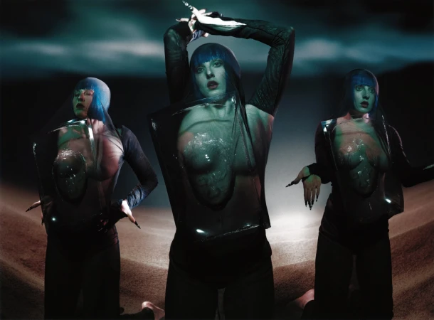 Three figures with blue hair and dark clothing pose against a dark background with ground, creating a surreal and futuristic aesthetic.