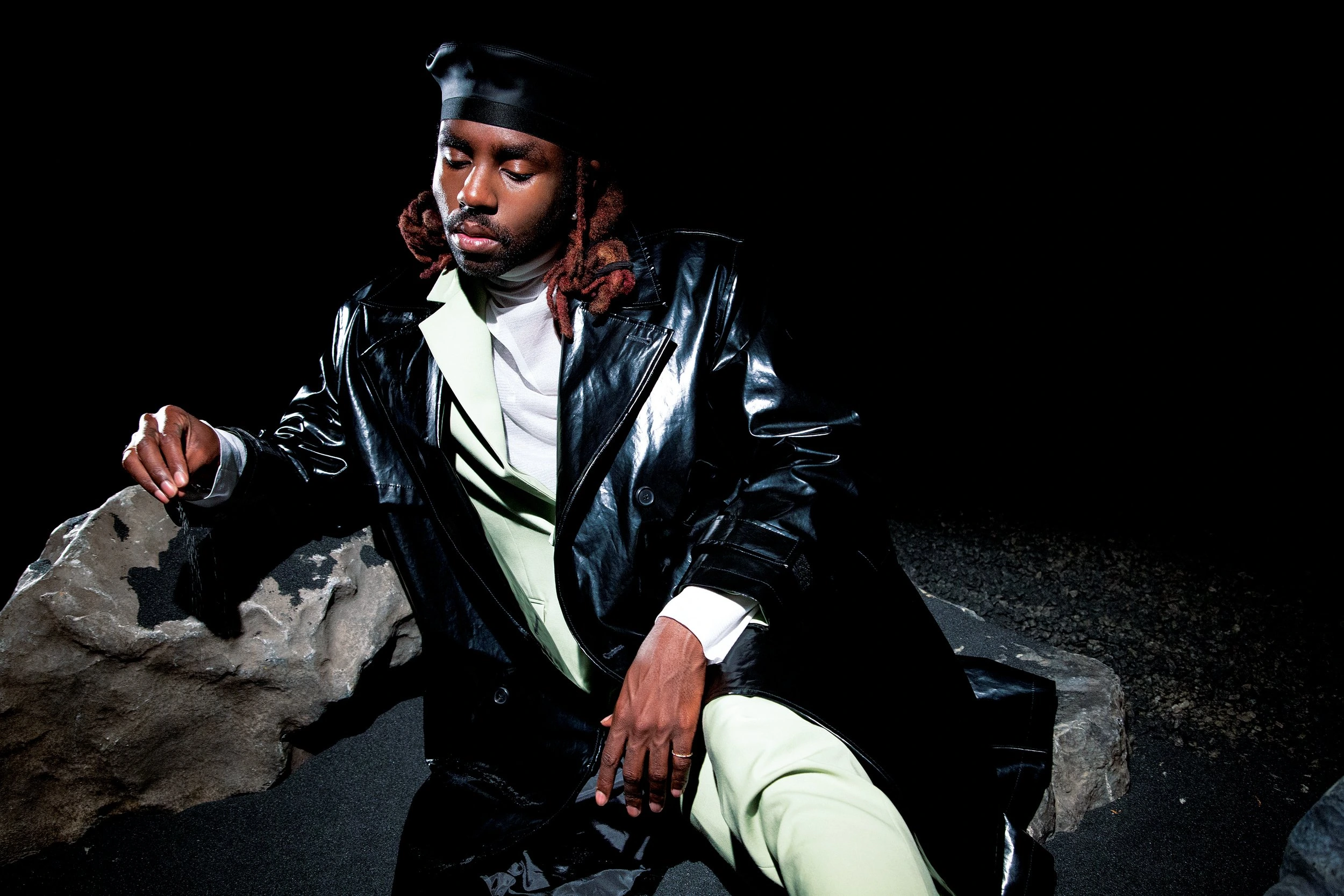 An adult male model poses on a rock in a black patent leather jacket and hat in a dimly lit setting, looking downward.