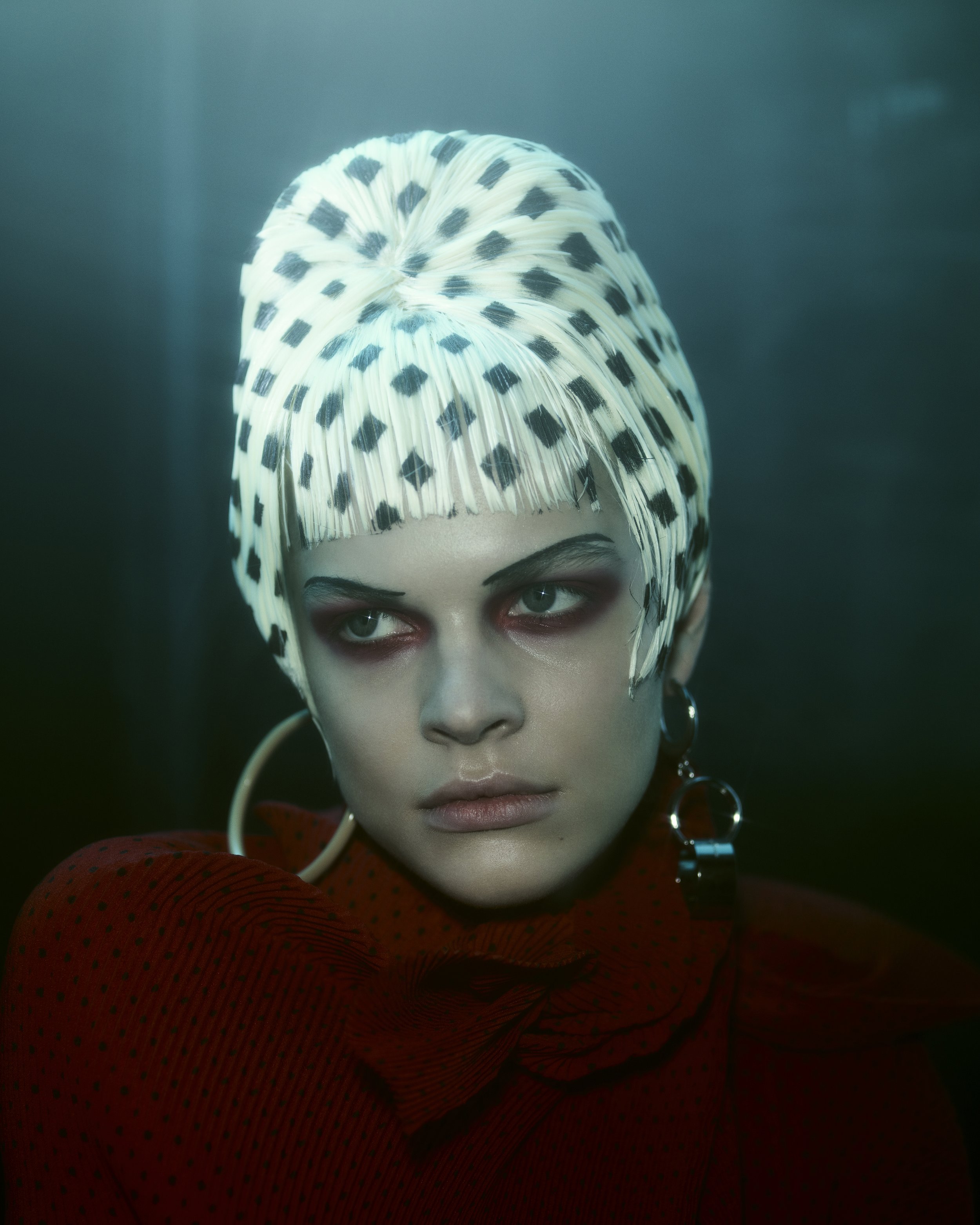 Close-up of a woman with fair skin, wearing a patterned white head covering, red top, and hoop earrings, set against a dark background.