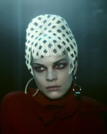 Close-up of a woman with fair skin, wearing a patterned white head covering, red top, and hoop earrings, set against a dark background.