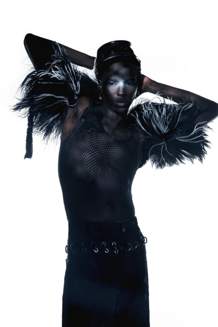 Striking fashion portrait of an androgynous figure with dark skin, wearing a textured top, elaborate shoulder pieces, and a black cap.