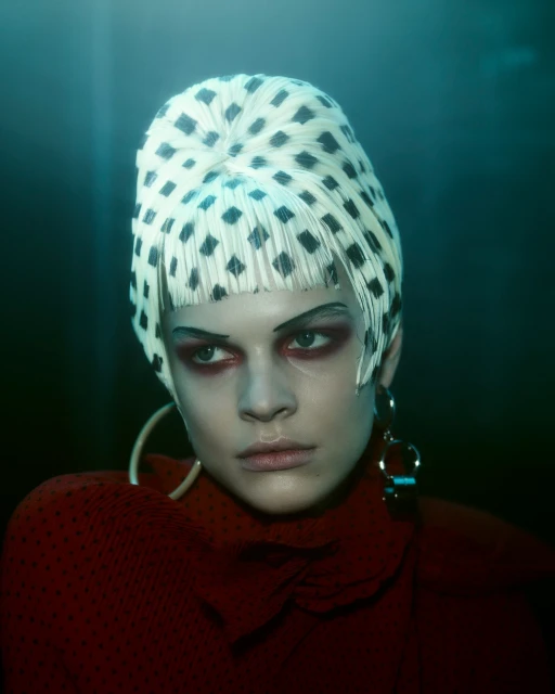 Close-up portrait of a model with a patterned white and black head covering, dramatic eye makeup, hoop earrings, and a dark red top.