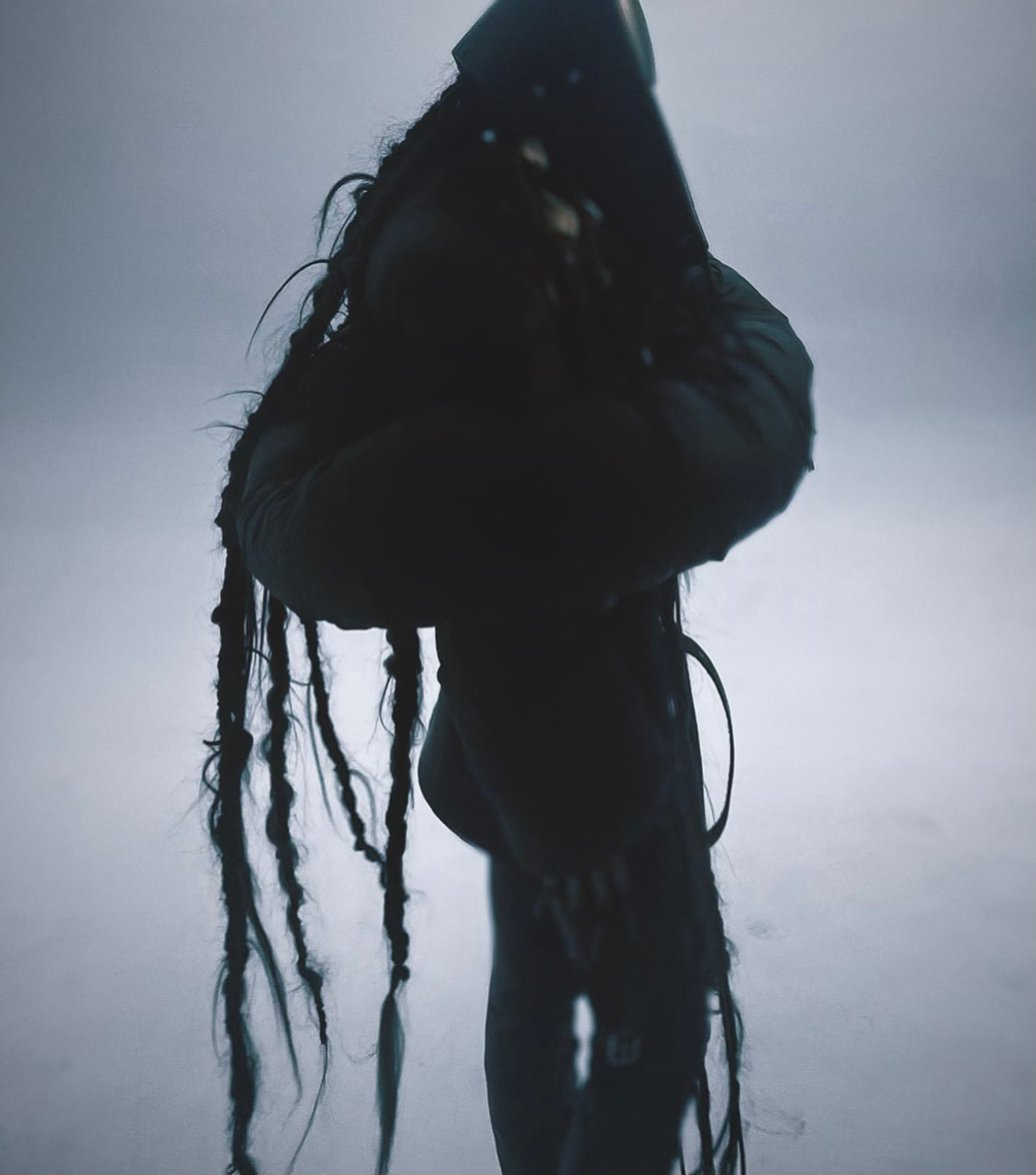 Monochromatic, mysterious image of a person with long braids wearing a dark hooded outfit, standing against a foggy, washed-out background.