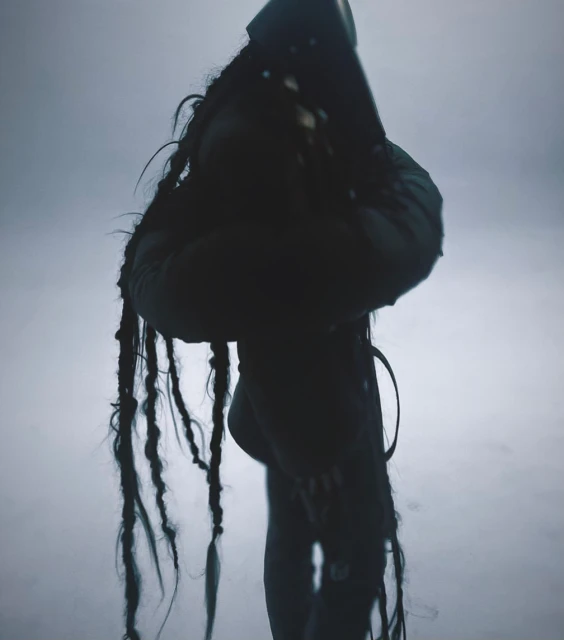 Monochromatic, mysterious image of a person with long braids wearing a dark hooded outfit, standing against a foggy, washed-out background.