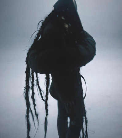 Monochromatic, mysterious image of a person with long braids wearing a dark hooded outfit, standing against a foggy, washed-out background.