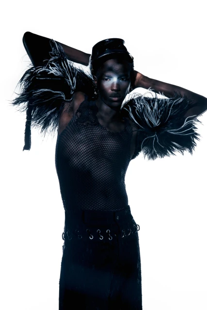 Dark-skinned model in black textured outfit with dramatic feathered shoulders poses in front of a bright white background.