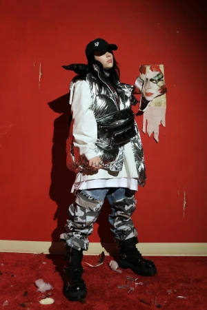 Billie Eilish wearing metallic puffer vest, camo cargo pants, and black cap holding a torn portrait against a red wall.