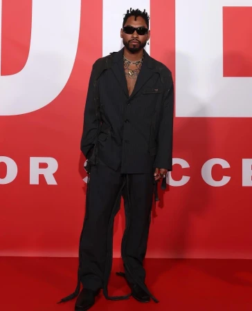 Man wearing dark sunglasses and dark grey suit standing in front of a bold red backdrop with white text