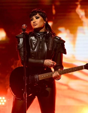 Singer Demi Lovato performs on stage with an electric guitar, wearing a black outfit, under bright stage lights at a music venue.