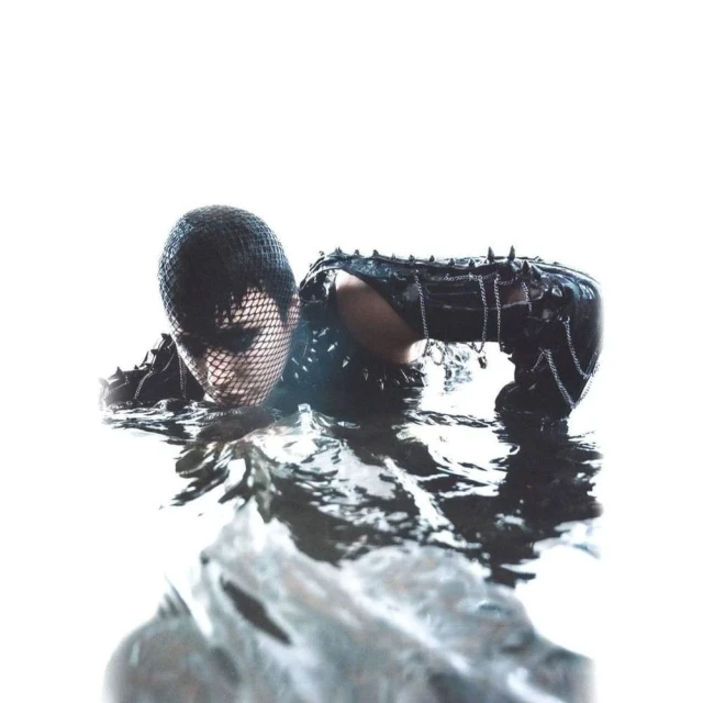 A woman with a net on her head emerges from dark water wearing a black, spiked leather outfit against a bright white background.