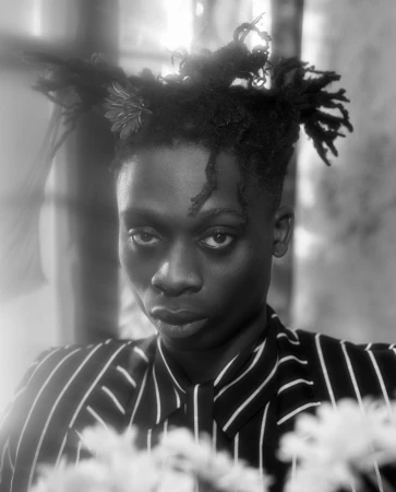 Monochrome portrait of a Black man with flowers styled in his dreadlocks, wearing a striped shirt.