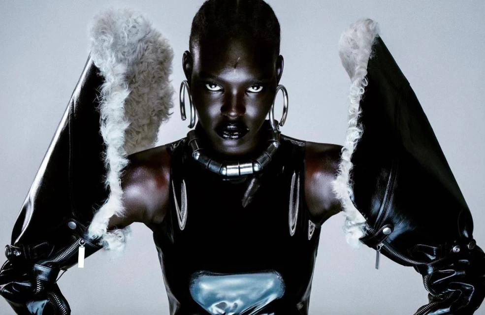 Headshot of dark-skinned adult woman in a black shiny latex top and black furry-trimmed sleeves looking intensely forward in a studio.