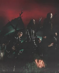 Group of four men in dark clothing posing in a surreal, moody landscape with dark twisted trees and a glowing red sky.