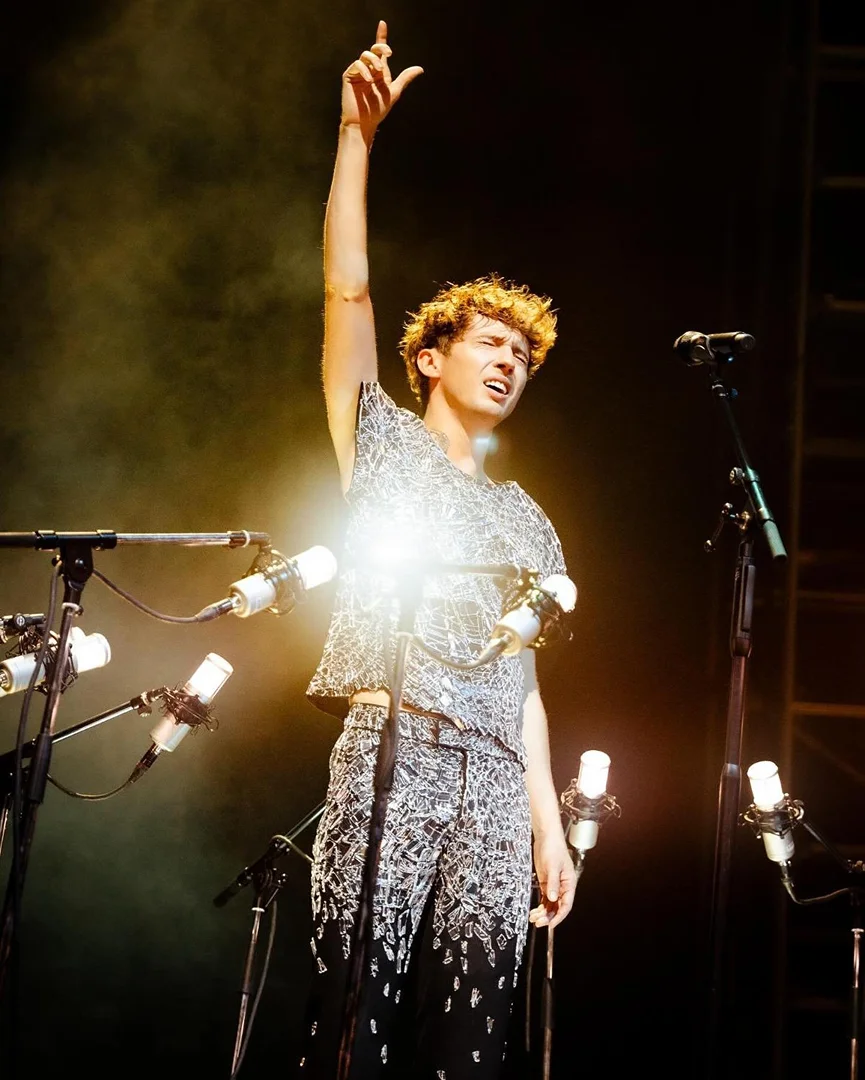Singer with curly hair performs on stage with multiple microphones, wearing a patterned shirt and pants with one arm raised.