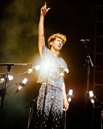 Singer with curly hair performs on stage with multiple microphones, wearing a patterned shirt and pants with one arm raised.