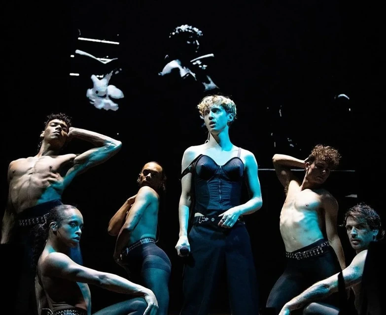 Performance on stage with a central performer in dark corset and pants surrounded by shirtless dancers, under black light and projected art.