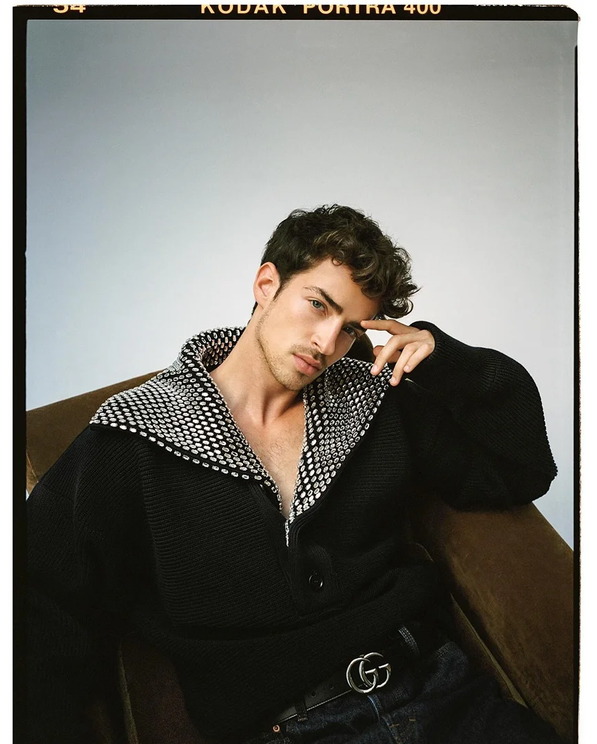 Portrait of a young adult man with curly hair, wearing a patterned black jacket and dark jeans, posing in a studio setting. 