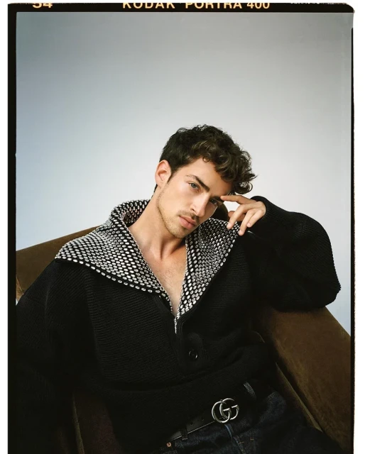 Portrait of a young adult man with curly hair, wearing a patterned black jacket and dark jeans, posing in a studio setting. 
