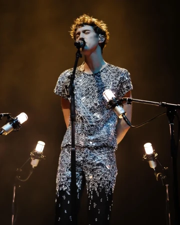 Singer performing live on stage with multiple microphones. He's wearing a shimmering outfit with dark hair, singing into a microphone.