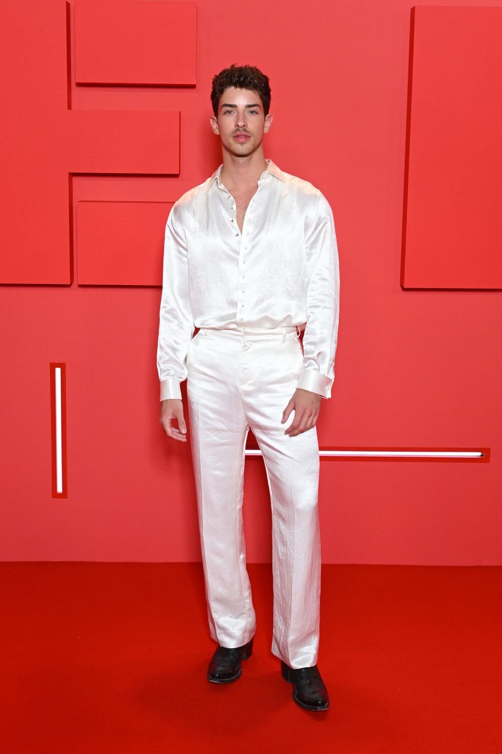 A dark-haired man in a white satin shirt and pants stands against a red backdrop with geometric shapes for a fashion event.