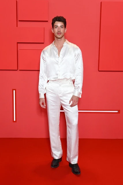 A dark-haired man in a white satin shirt and pants stands against a red backdrop with geometric shapes for a fashion event.