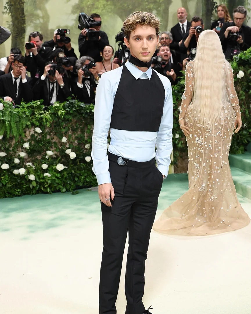 Young man in formal wear poses at an event with photographers in the background and a woman in a long gown walking away.
