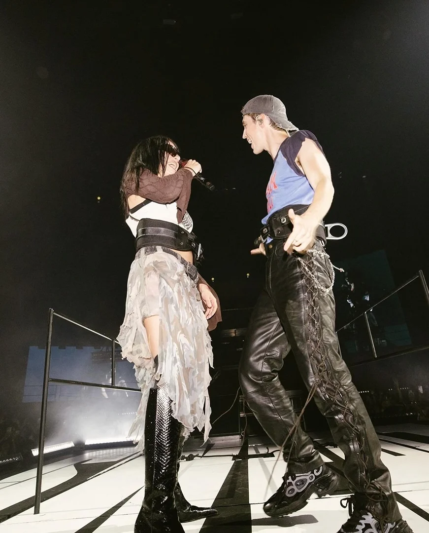 Two musicians, a male and female, perform on stage during a concert, captured from a low angle with stage lighting.