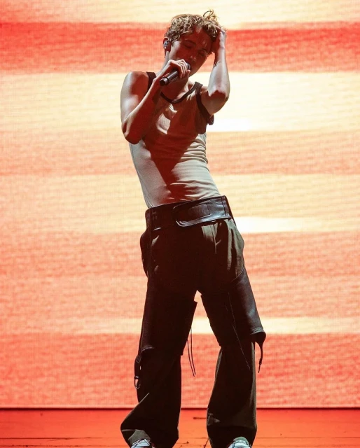 Male singer performs on stage wearing a tank top and baggy pants with a microphone in hand, set against a red and orange background.