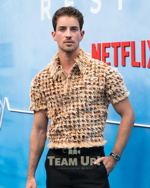 Adult male poses at an event. He wears a unique beige-brown textured shirt and black pants against a light blue backdrop.