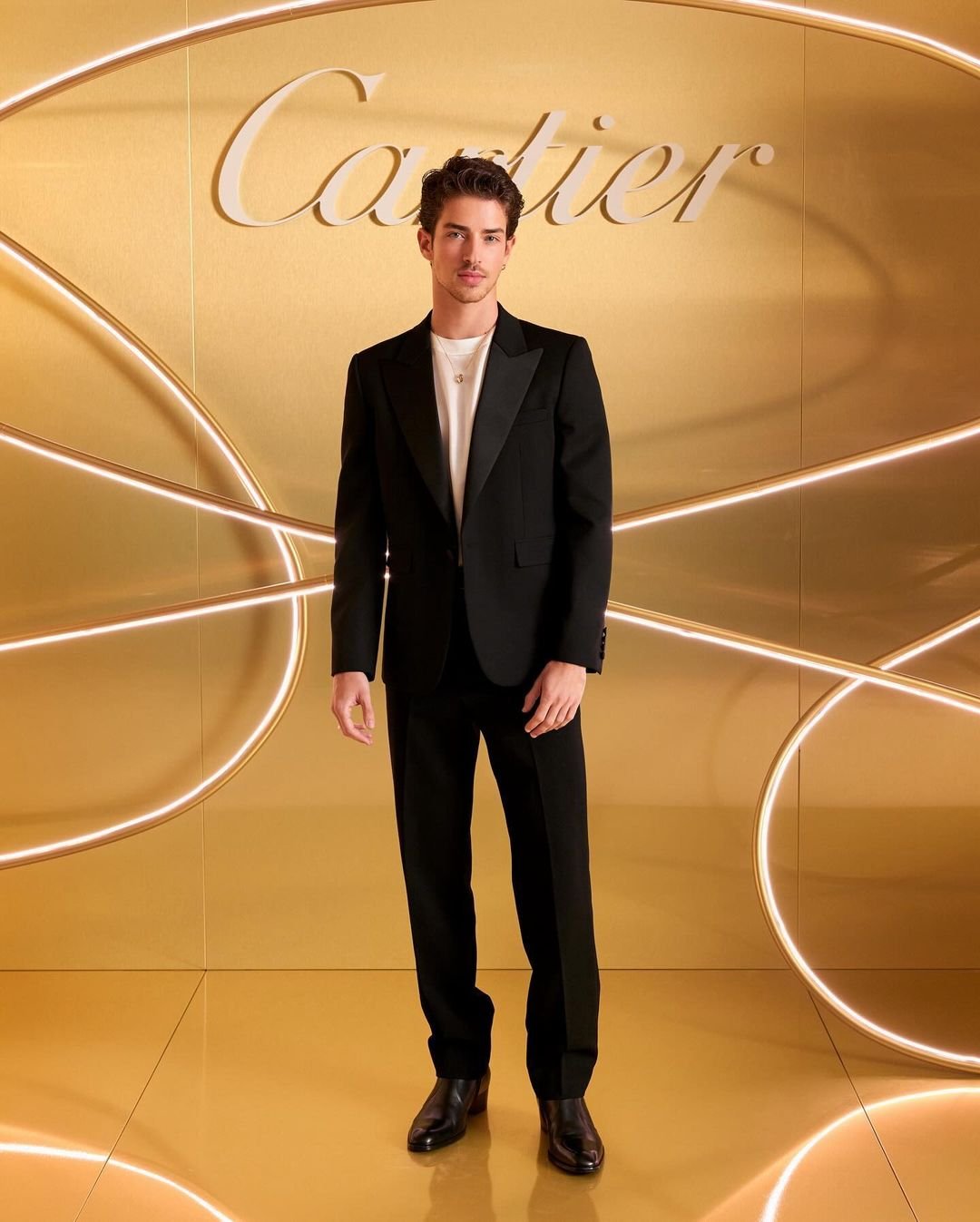 Portrait of a young adult man in a black tuxedo suit posing in front of a gilded wall with Cartier logo and curved neon lighting.