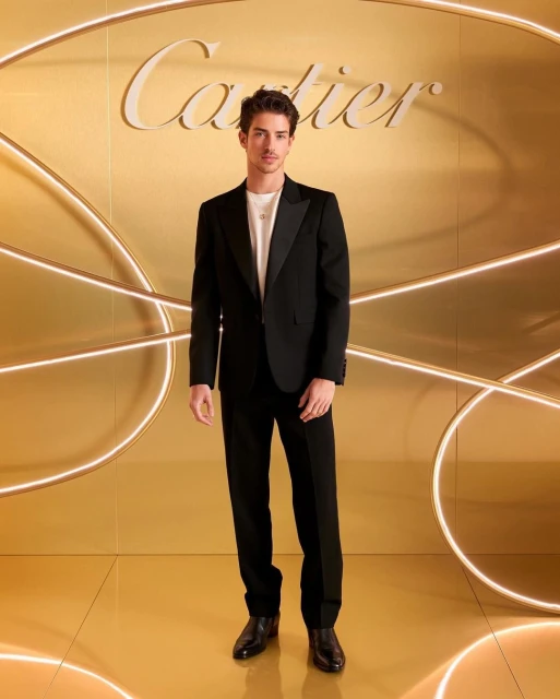 Portrait of a young adult man in a black tuxedo suit posing in front of a gilded wall with Cartier logo and curved neon lighting.