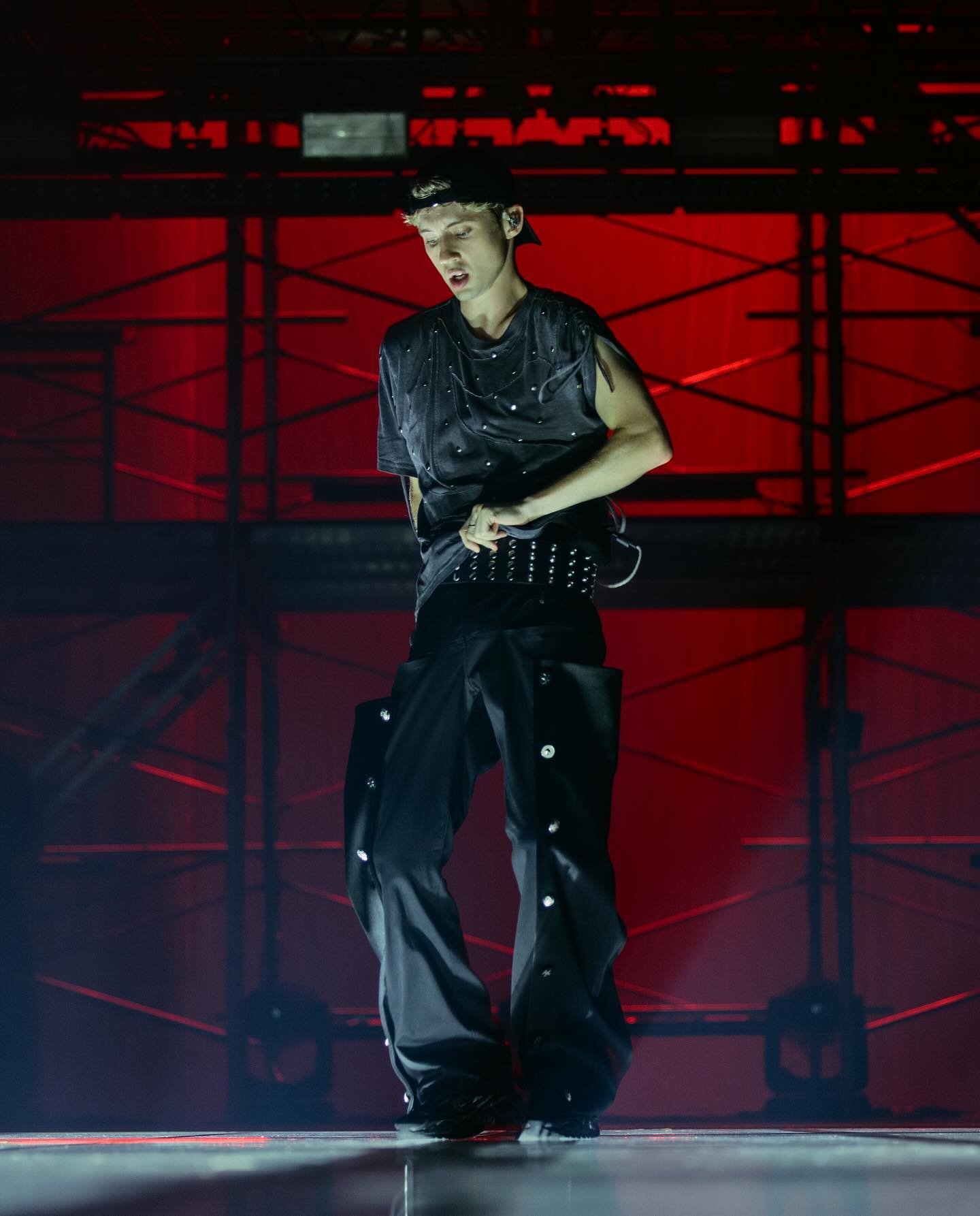 Singer performing on stage in a black top, pants, and cap under red stage lights, with a focused facial expression.